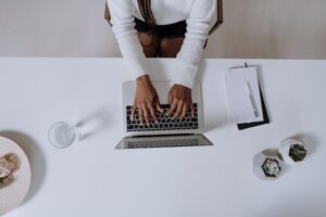 A woman in a white shirt typing on a laptop, a glass of water, and a notebook next to her on the table.